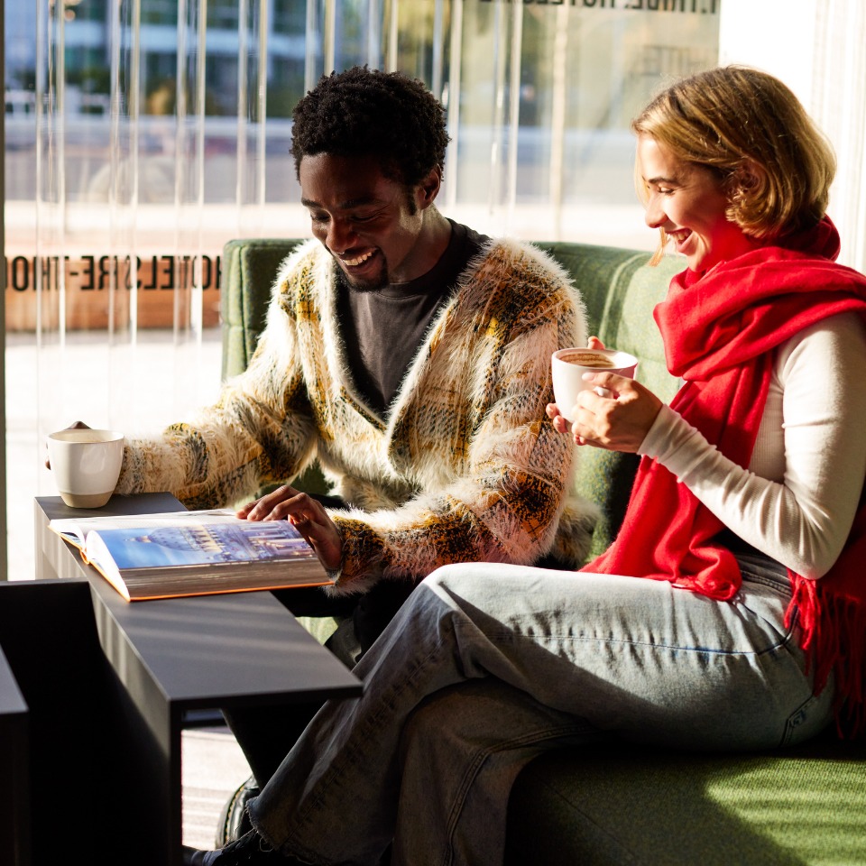 Two people engaged in conversation over drinks, with sunlight streaming through large windows.