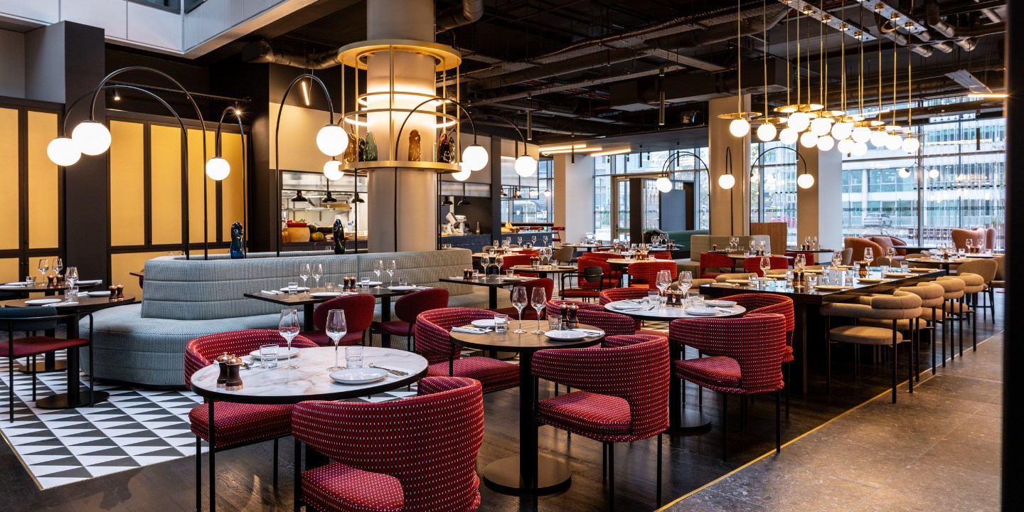 Modern restaurant interior with stylish red and grey seating, elegant tables set for dining, and bright overhead lighting.