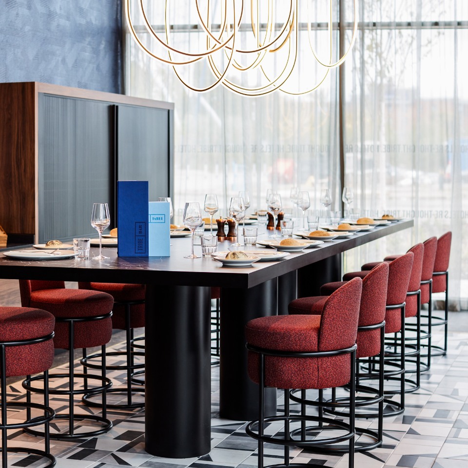 A long black table in a restaurant with floor to ceiling windows. The tables are set.