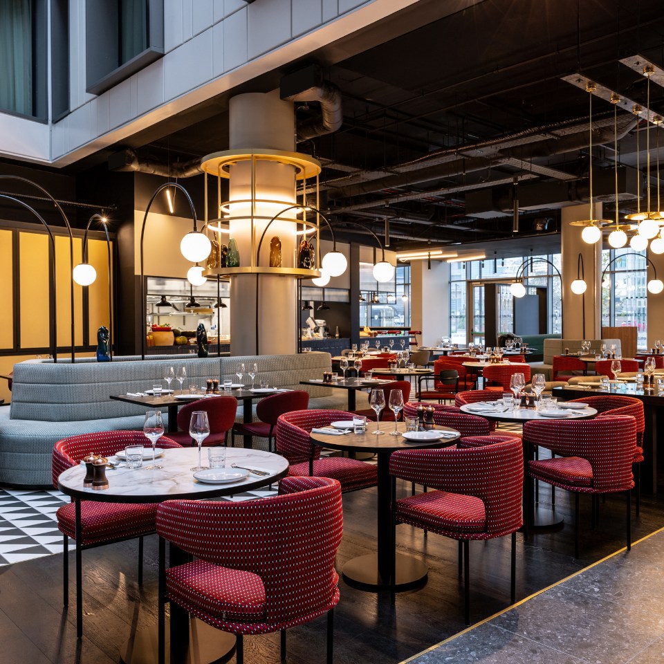 Modern restaurant interior with stylish red and grey seating, elegant tables set for dining, and bright overhead lighting.