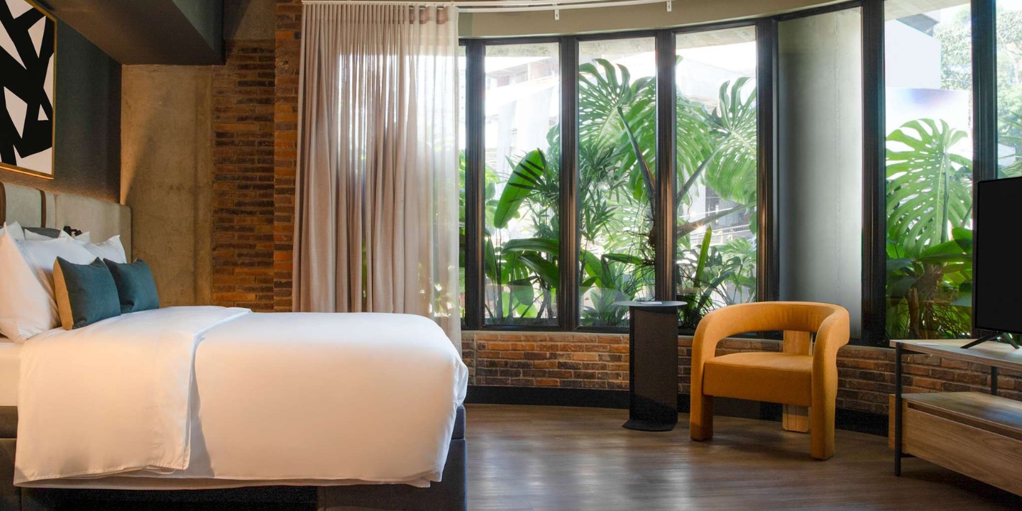 Modern hotel room with a plush bed, vibrant plants outside large windows, a cozy orange chair, and warm wood accents.