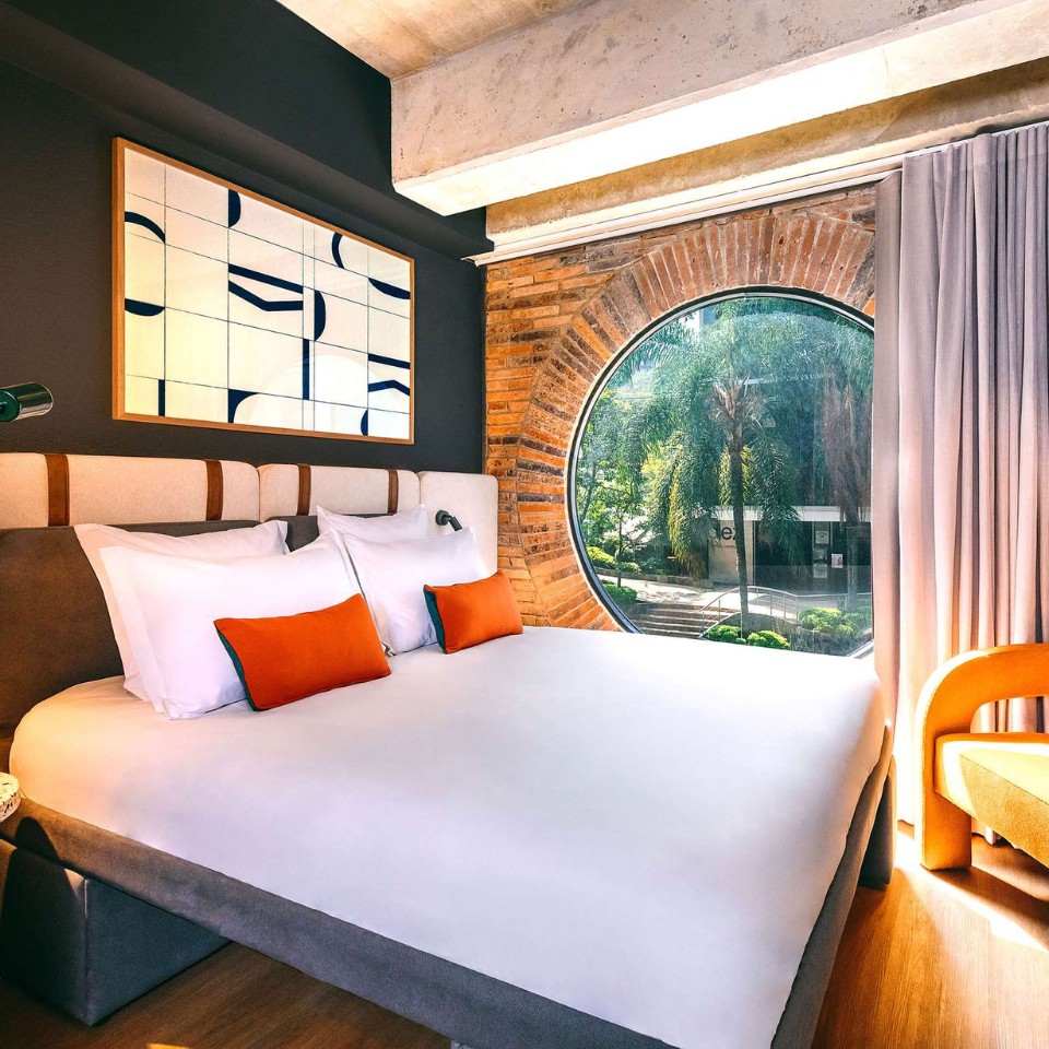Cozy hotel room featuring a circular window, modern decor, white bedding, and orange accent pillows, with greenery visible outside.