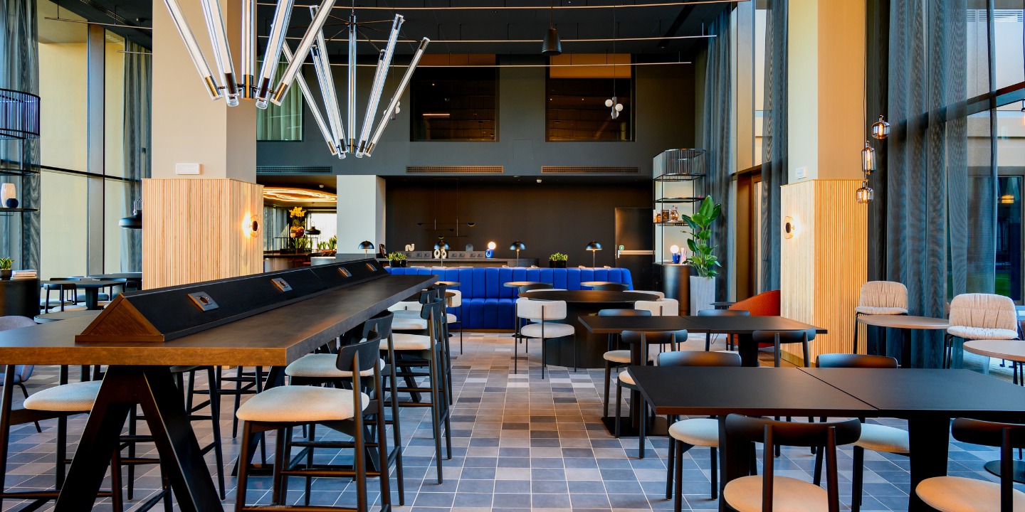 Modern dining area with sleek wooden tables, stylish chairs, and striking pendant lights, complemented by large windows and greenery.