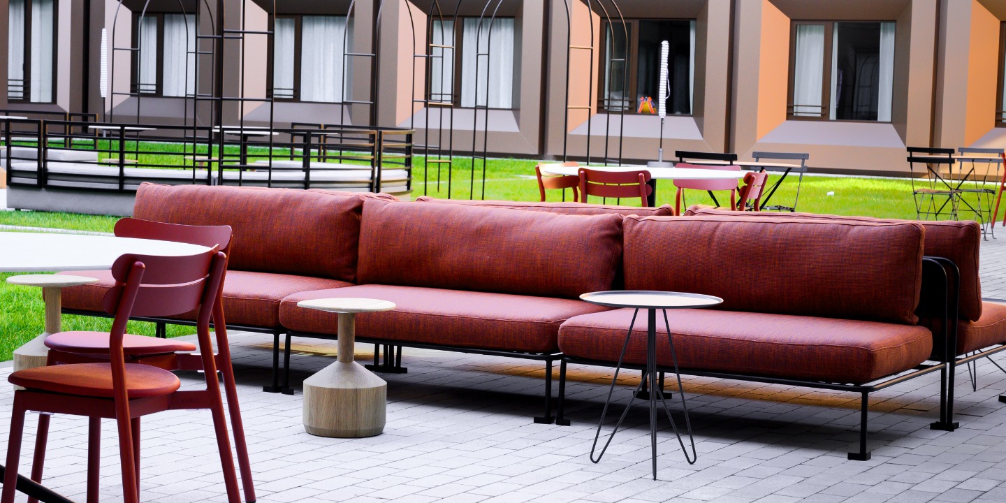 A modern outdoor seating area features a rust-colored sofa, minimalist tables, and red chairs, surrounded by green grass and contemporary buildings.