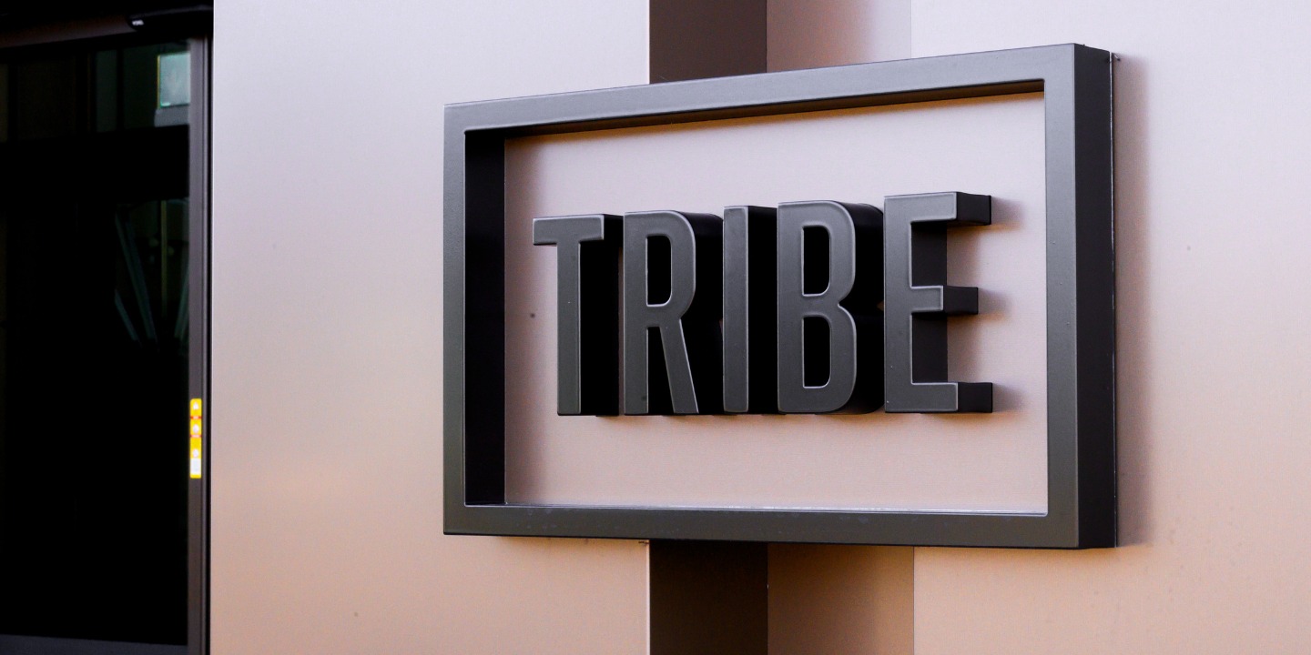 Close-up of a modern sign displaying the word "TRIBE" in bold black letters framed against a light-colored wall.