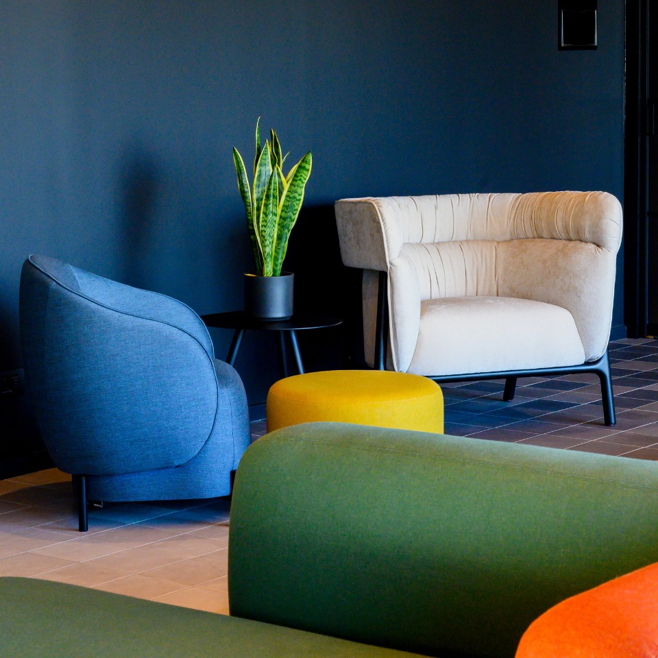 A modern lounge area featuring two stylish chairs, a round yellow ottoman, a small table with a plant, and a dark wall backdrop.