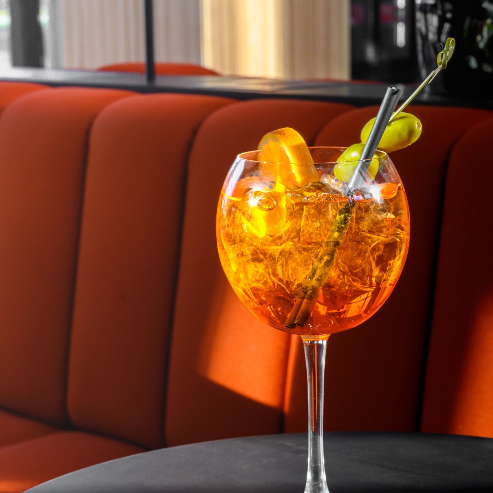 A large, bright orange cocktail with ice, garnished with an orange slice and olives, sits on a black table beside an orange couch.