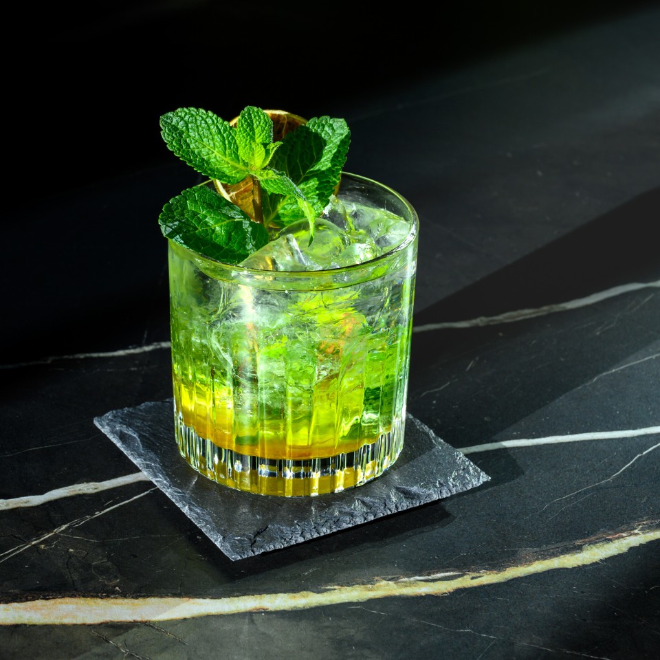 A refreshing cocktail sits on a slate coaster, featuring mint leaves and ice, with vibrant green and yellow layers, against a dark backdrop.