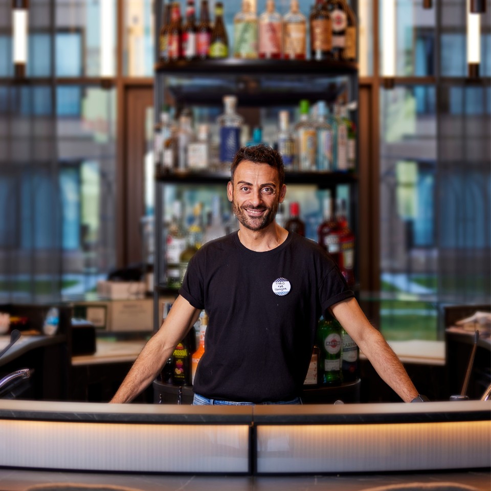 A bartender in a black shirt stands behind a well-stocked bar, showcasing various bottles of liquor and drinks.