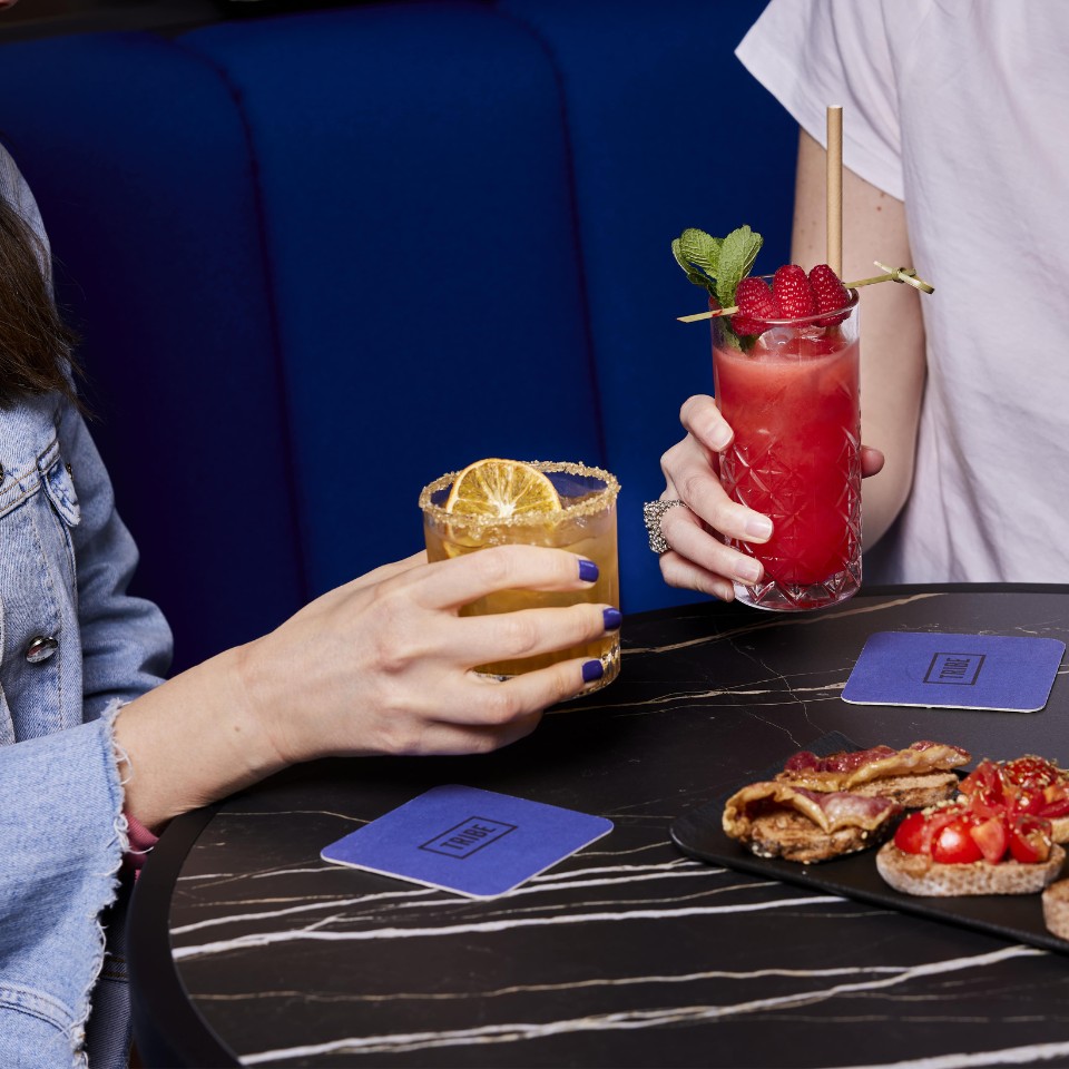 Two hands hold colorful cocktails, with a platter of appetizers on a stylish table and vibrant blue seating in the background.