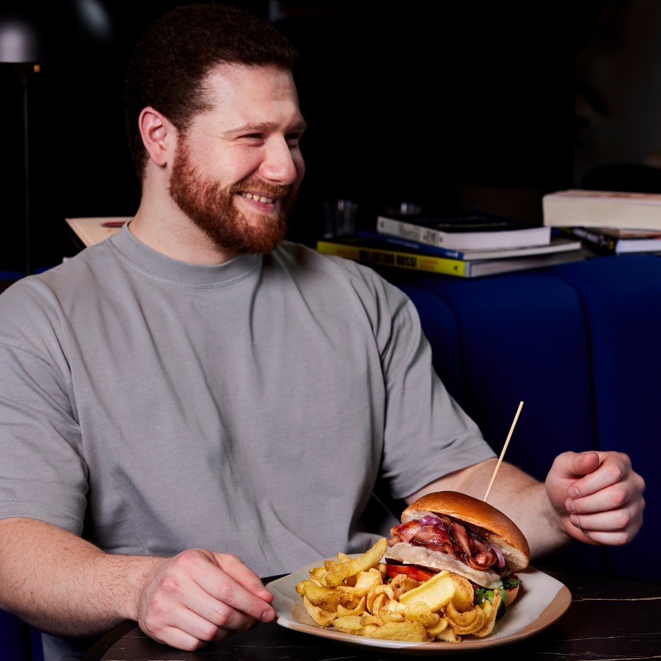 A person sits at a table holding a plate with a large burger topped with bacon, accompanied by crispy potato chips.