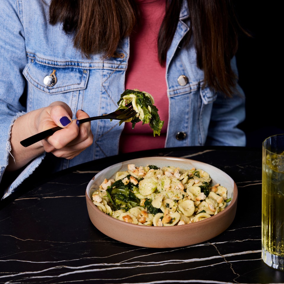 A woman holds a fork with pasta and greens above a bowl of orecchiette, with a drink beside her on a dark marble table.