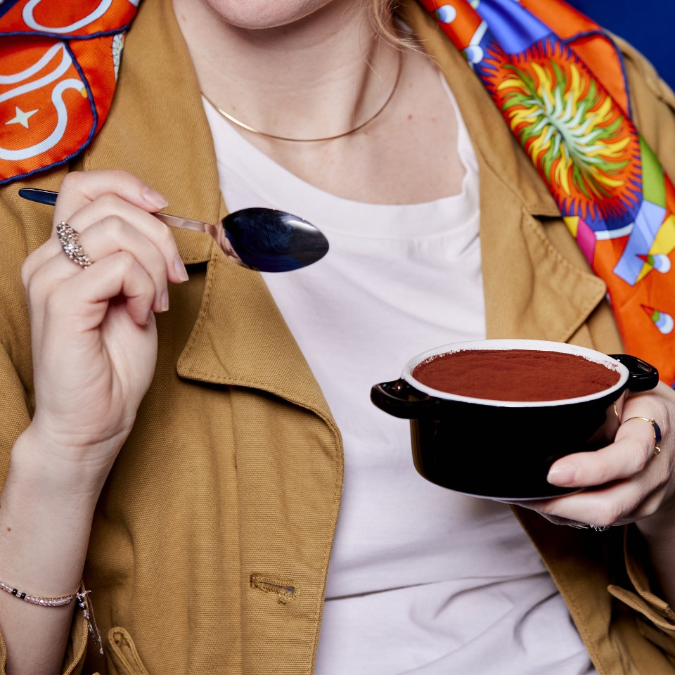 A person in a tan jacket holds a small black dish of dessert, ready to take a bite with a spoon, wearing colorful accessories.