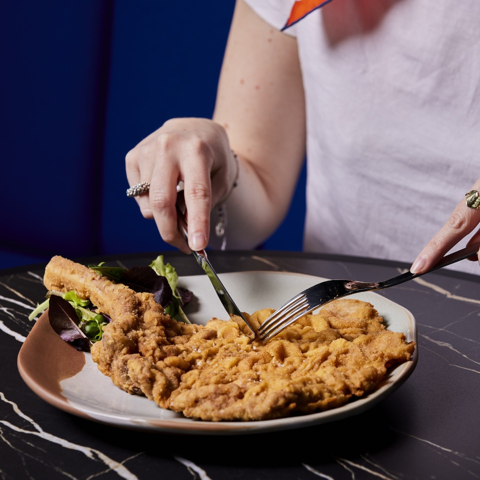 A person using a fork and knife to cut into a large fried pork chop on a plate, accompanied by a side salad.