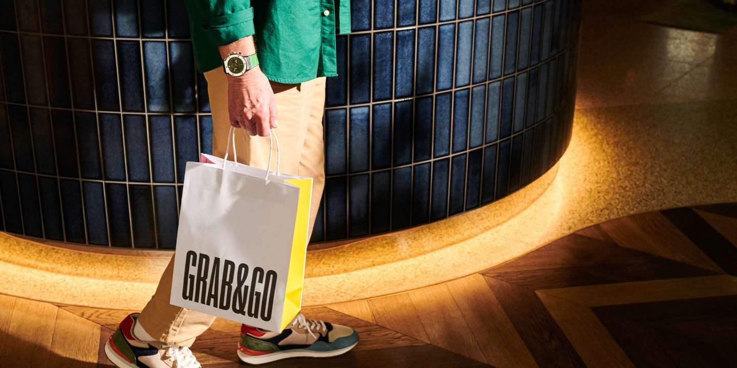A person in a green shirt and tan pants carries a "GRAB&GO" shopping bag while walking on a stylish wood floor.