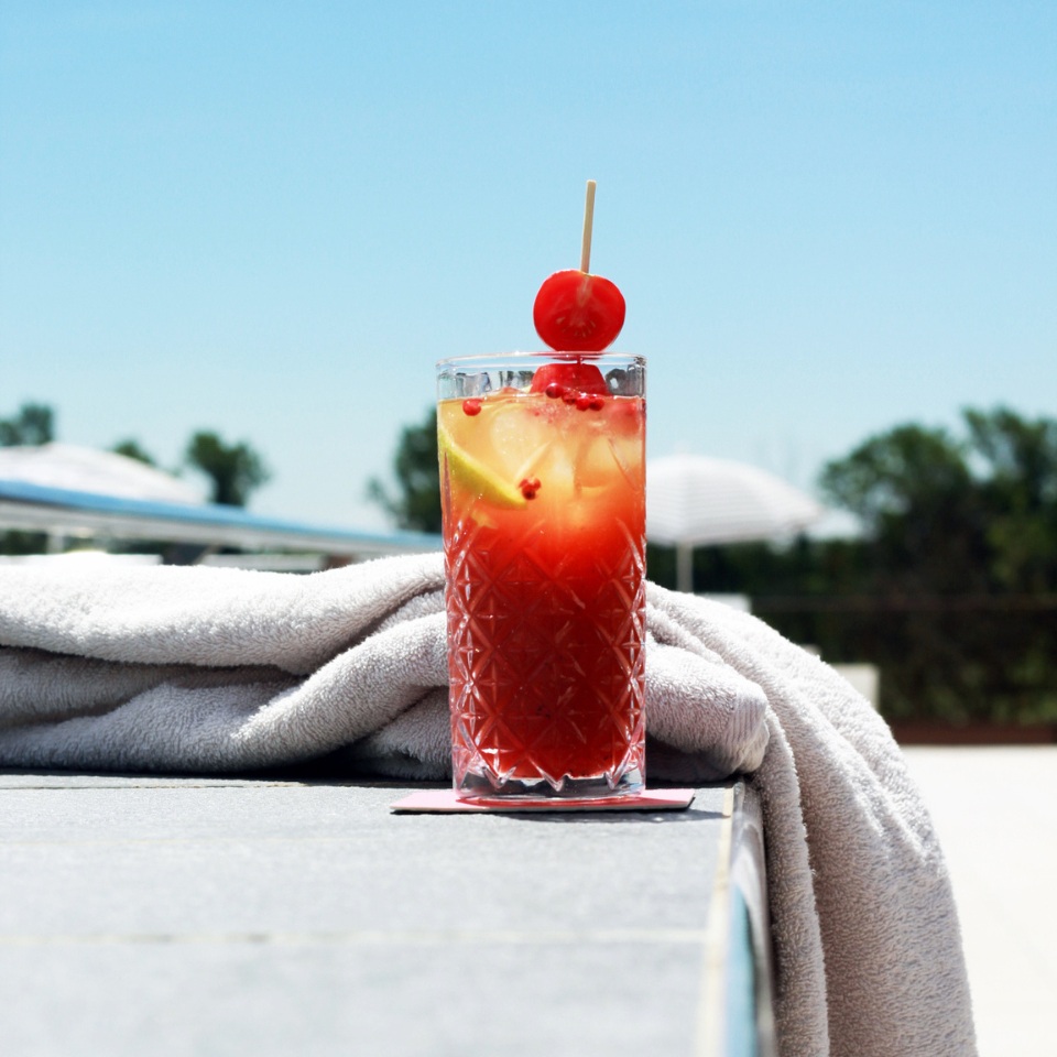 A refreshing red cocktail garnished with a cherry and lemon slice, resting on a stone surface beside a white towel and bright blue sky.