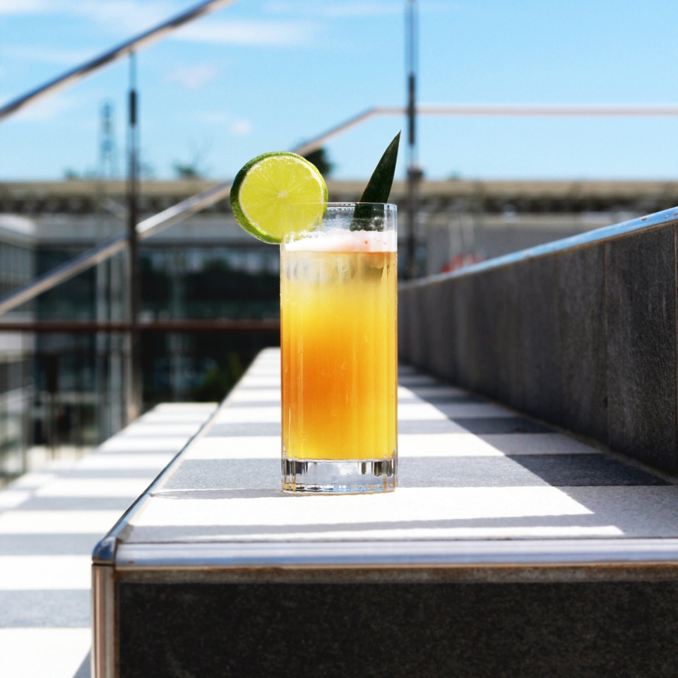 A tall glass of a refreshing orange cocktail garnished with a lime slice and a green leaf, sitting on a stylish staircase in bright sunlight.