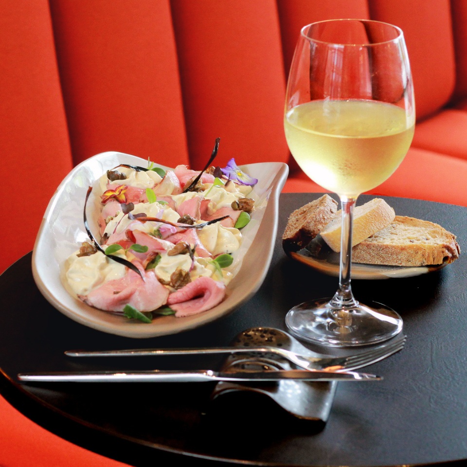 A plate of colorful salad with pink meat, creamy sauce, and edible flowers, paired with a glass of white wine and bread on a table.