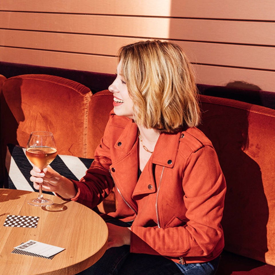 A woman sits at a table with a glass of wine, wearing an orange jacket, against a backdrop of warm-toned upholstery.