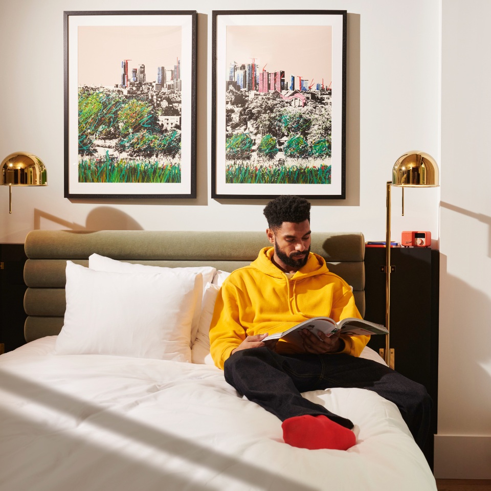 A person in a yellow hoodie sits on a bed, reading a book, with two framed artworks of a city skyline on the wall behind them.