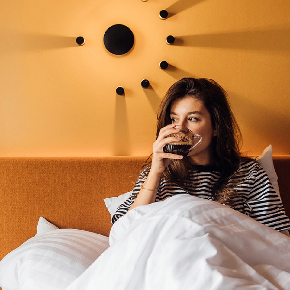 A person sits up in bed, wrapped in white blankets, against a warm orange wall with a modern lamp decor.