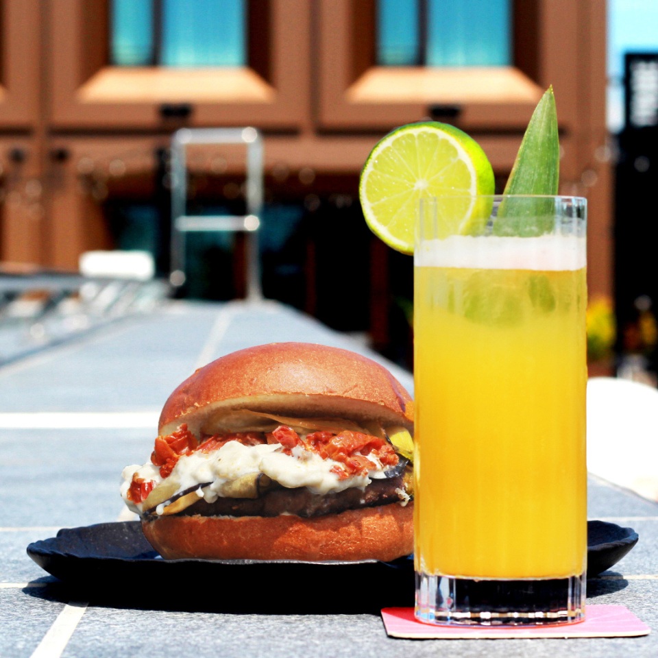 A juicy burger with toppings sits on a black plate next to a tall glass of orange drink garnished with lime, set on a sunny table.