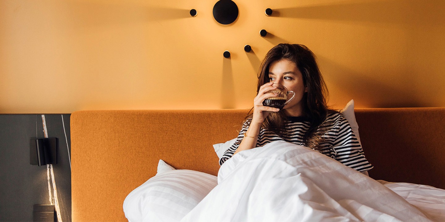 A person sits up in bed, wrapped in white blankets, against a warm orange wall with a modern lamp decor.