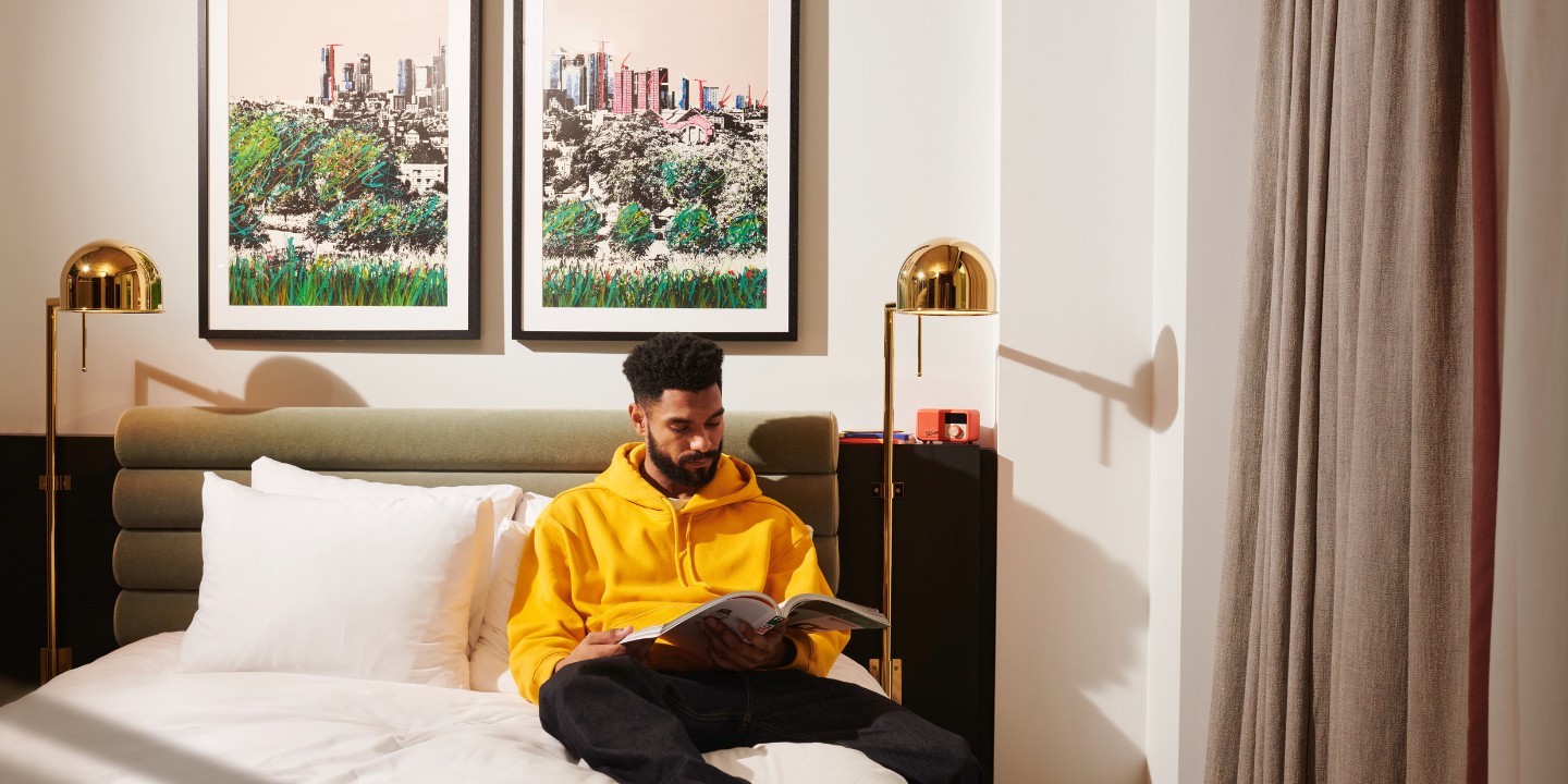 A person in a yellow hoodie sits on a bed, reading a book, with two framed artworks of a city skyline on the wall behind them.