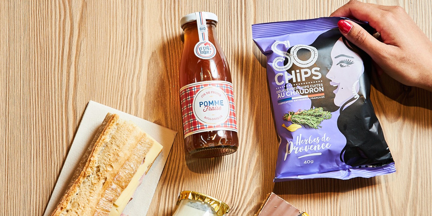 A wooden table with a baguette sandwich, a bottle of apple soda, a jar of dessert, chips, and a chocolate snack.