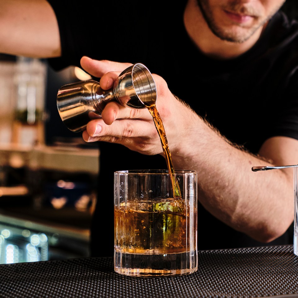 A bartender, focused and intent, pours amber liquid from a jigger into a glass on a bar counter, conveying a mood of skill and precision.