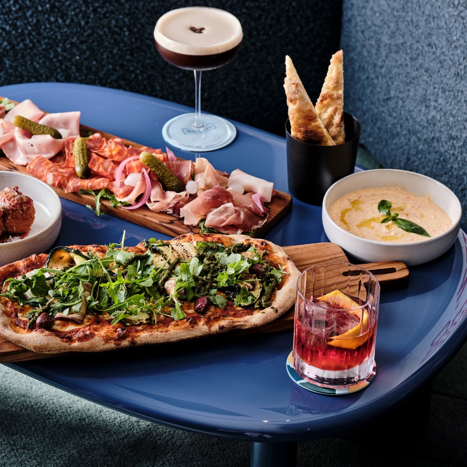 A vibrant spread on a blue table includes a pizza topped with greens, a charcuterie board, cocktails, breadsticks, and a creamy dip, creating a lively and inviting dining atmosphere.