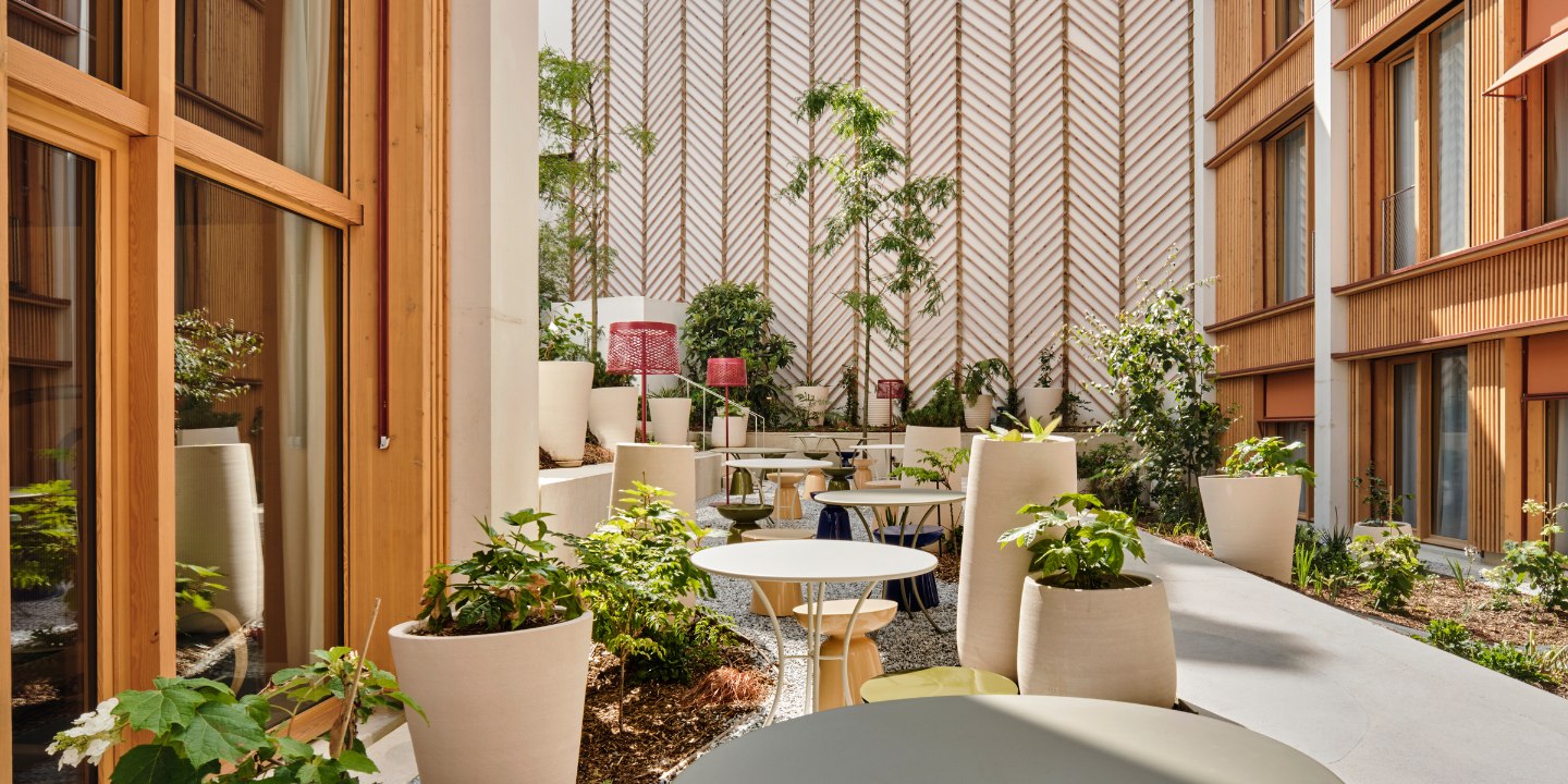 A serene patio with circular tables, lush potted plants, and cosy chairs is framed by warm wood-paneled walls, offering a tranquil, inviting outdoor space.