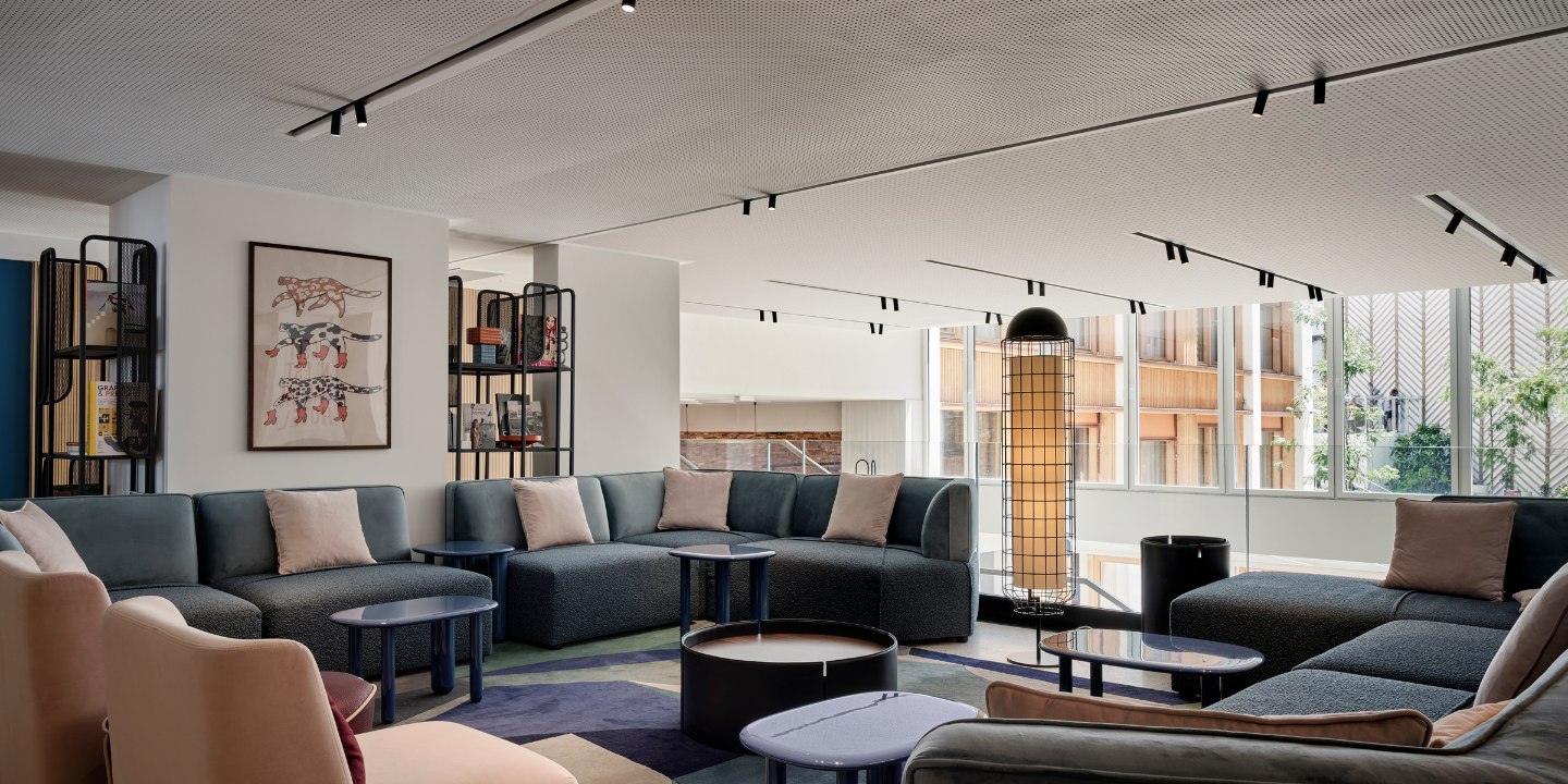 Modern lounge with blue and beige sofas, pink cushions, and round tables. Large windows, grid-patterned lamp, art on walls, and a cozy atmosphere.