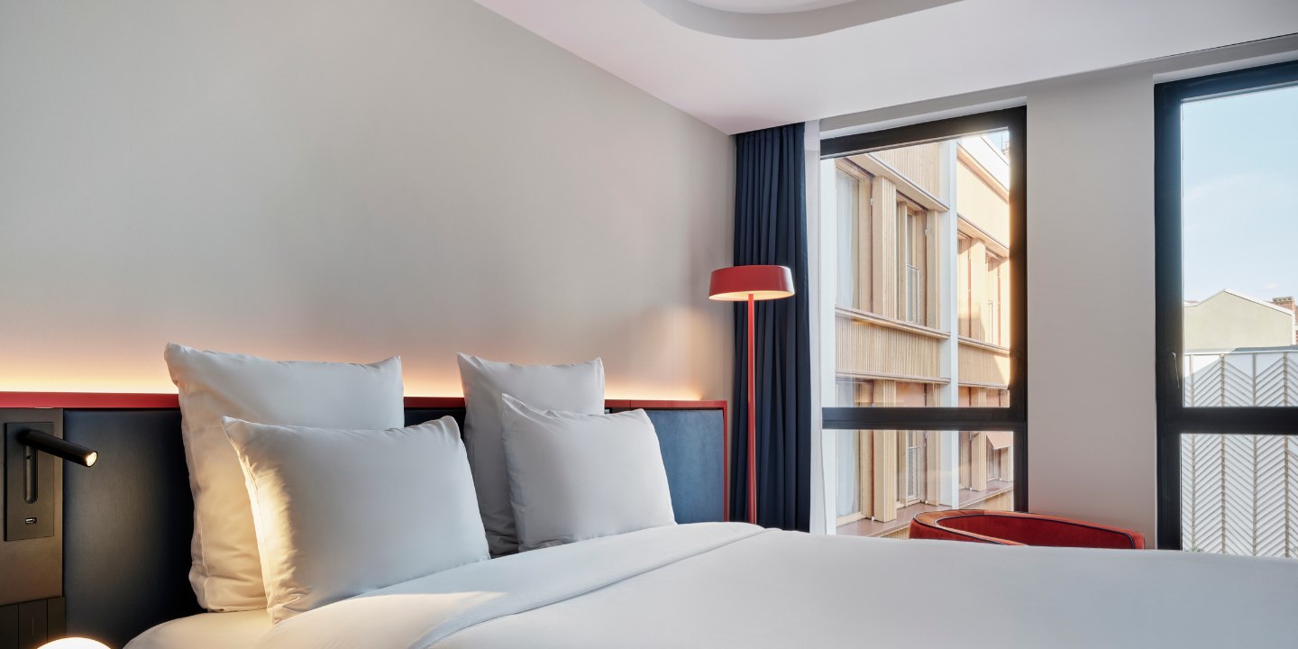 Modern hotel room with a neatly made bed, two plush pillows, sleek black bedside lamp, red floor lamp, large windows, and warm, inviting tones.
