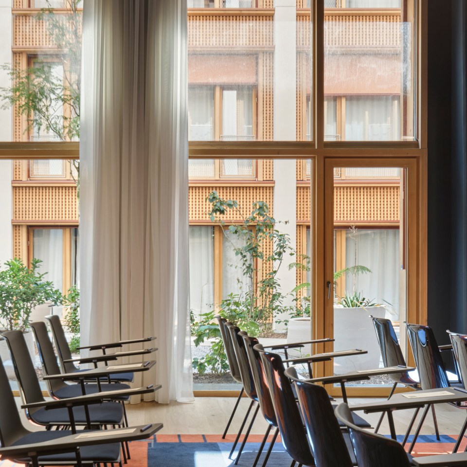 Sunlit room with rows of empty chairs facing large windows, sheer curtains, and a view of a building exterior with plants outside, creating a calm ambiance.