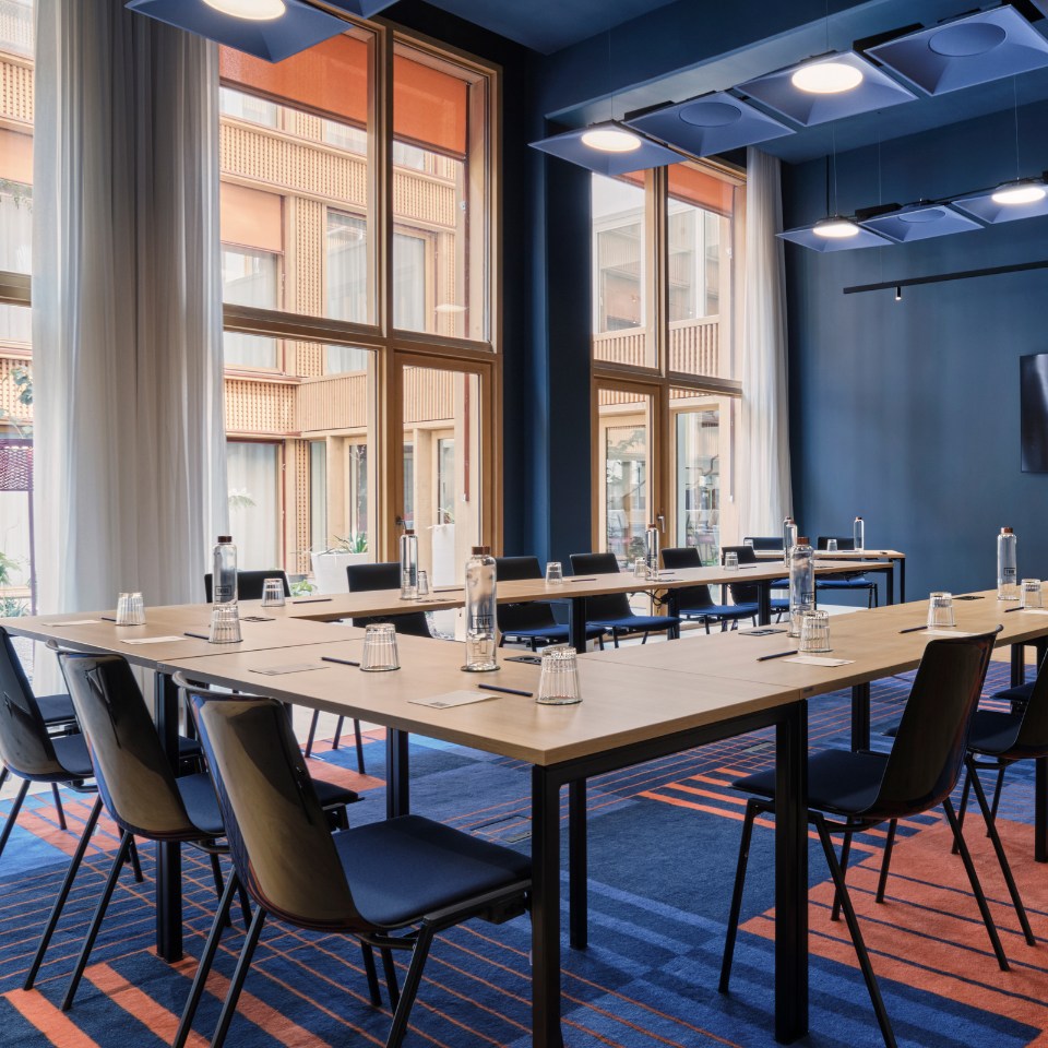 A modern, well-lit conference room with large windows, blue walls, and orange-striped carpet. Tables are arranged in a U-shape with chairs and water bottles.