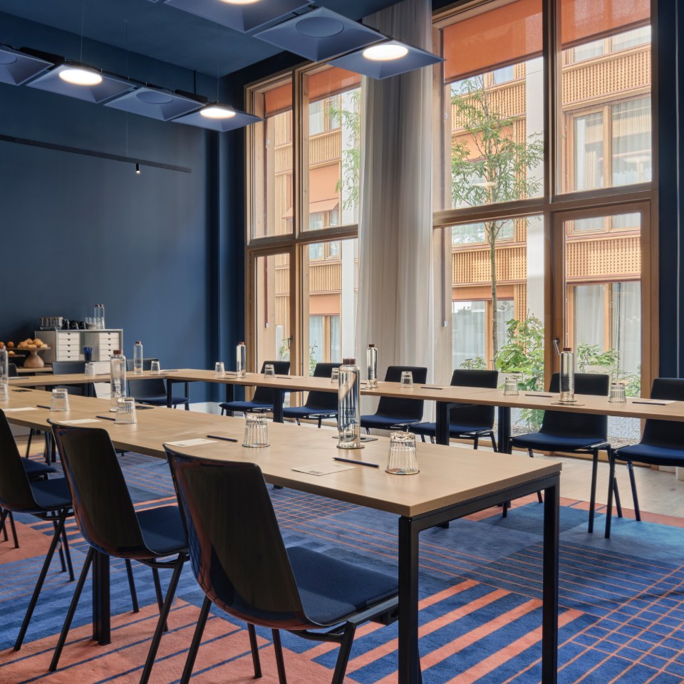 Modern conference room with long tables and navy chairs, set with water bottles and glasses. Large windows reveal a view of an orange building and greenery.