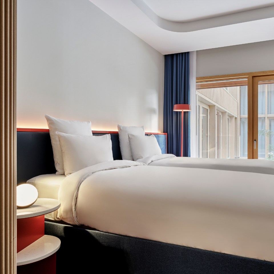 Modern hotel room with two neatly made beds featuring plush white bedding. Warm lighting and large windows add a cozy, inviting atmosphere.