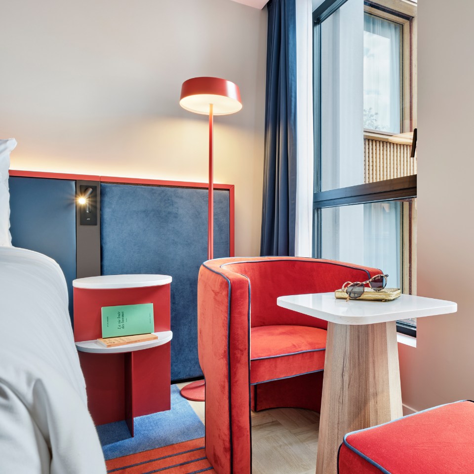 Modern hotel room with vibrant red and blue decor. Features include a red armchair, stylish floor lamp, small white table with sunglasses, and a large window.