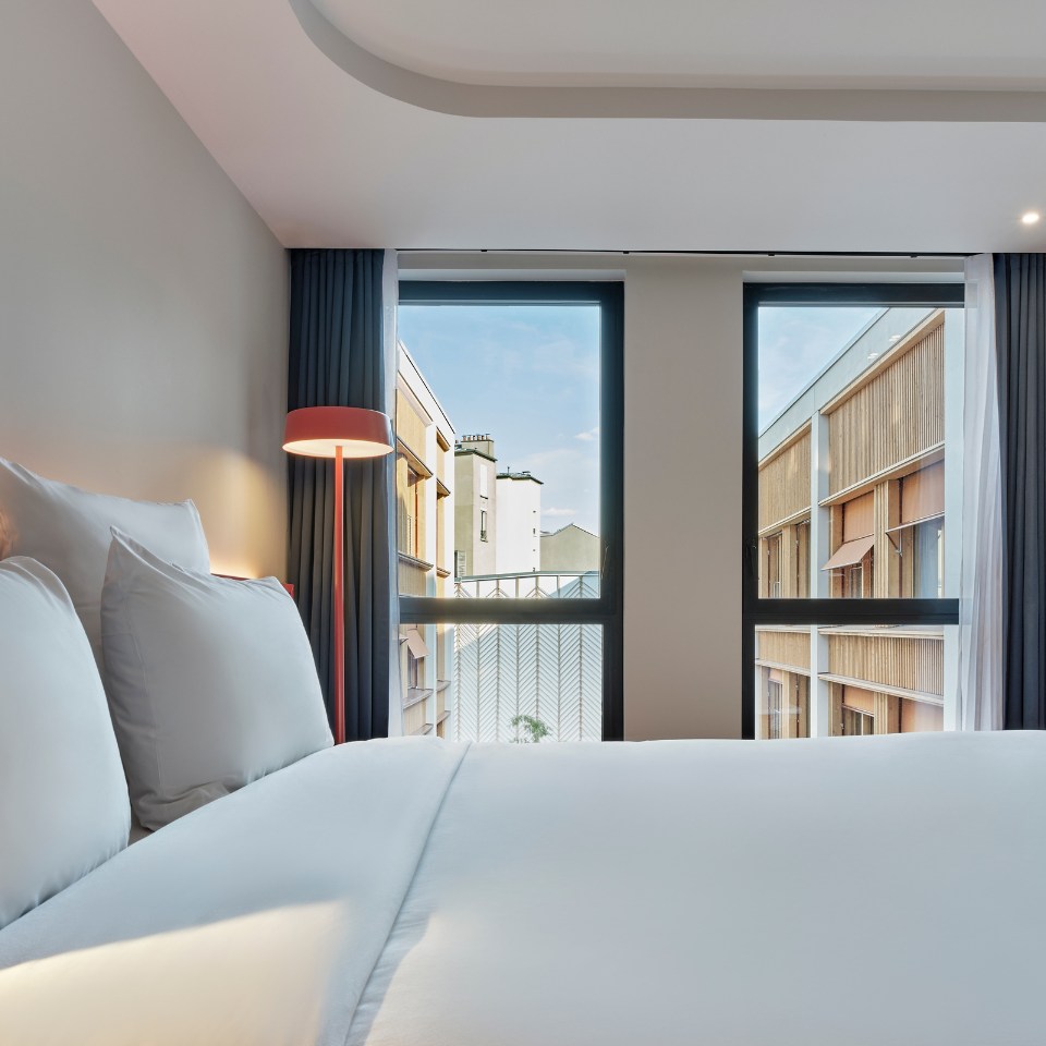 A modern hotel room featuring a neatly made bed with white linens, a tall red floor lamp, and large windows offering a view of neighboring buildings under a blue sky.