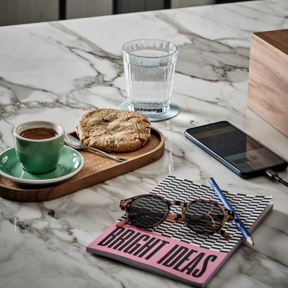 A marble table holds a coffee in a green cup and a cookie on a wooden tray, a water glass, a phone, sunglasses, and a "Bright Ideas" notebook with a pencil.