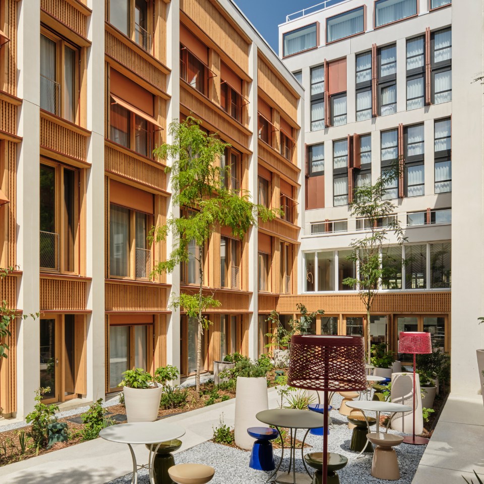 Modern courtyard with warm-toned residential buildings and a vibrant garden. Features colorful tables and chairs, with a bright, inviting atmosphere.