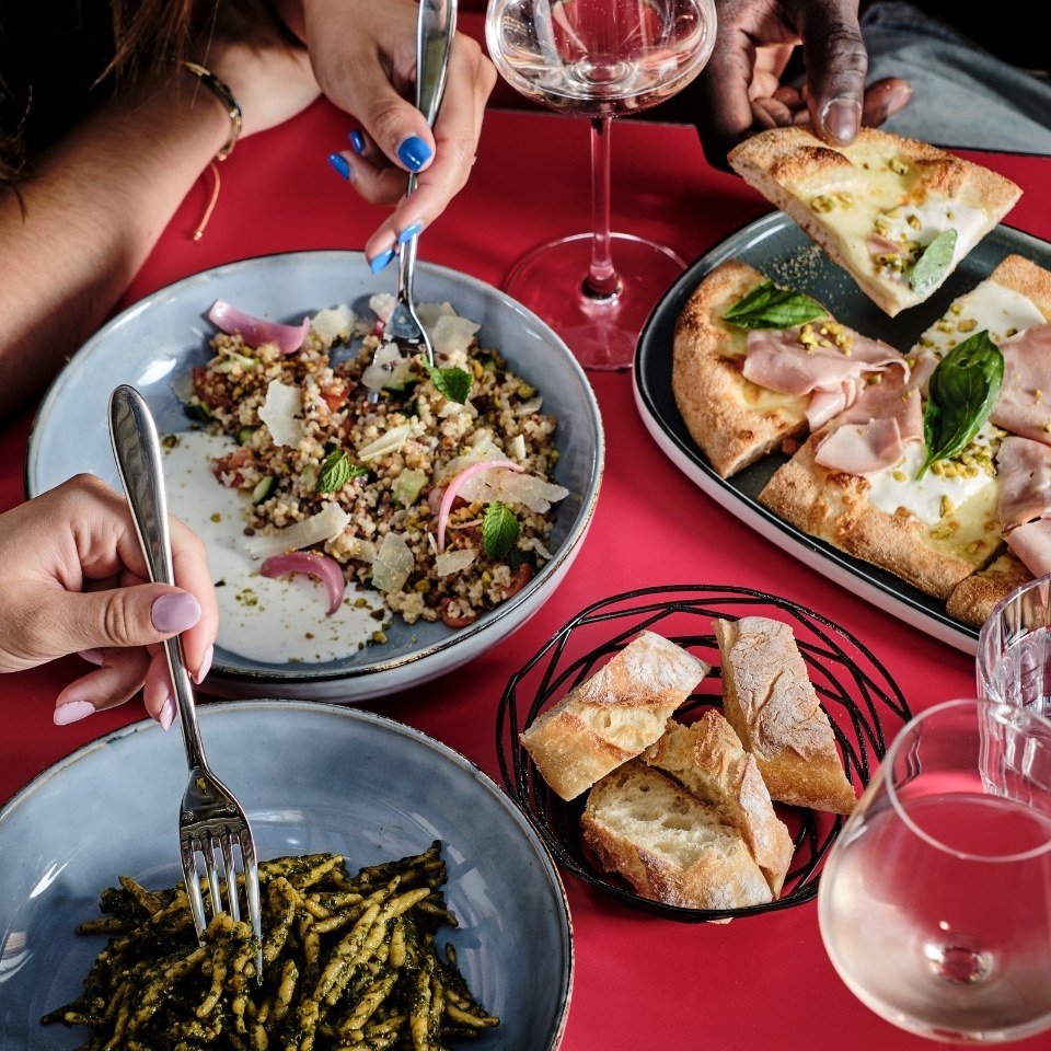 A vibrant table setting with hands reaching for food. Dishes include couscous salad, pasta, pizza, bread, and wine, creating a lively, shared dining experience.