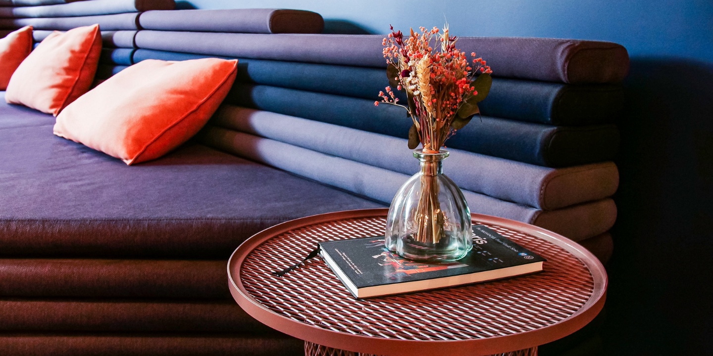A small red table with a book and a vase of dried flowers, in the backgournd is a large purple couch with orange pillows