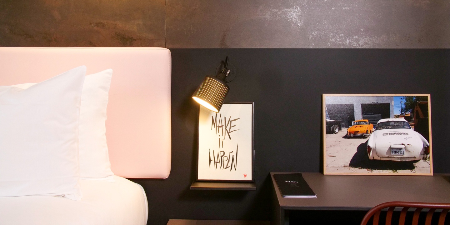 A bed with a pink headboard, to the right is a small desk with a framed picture of a car