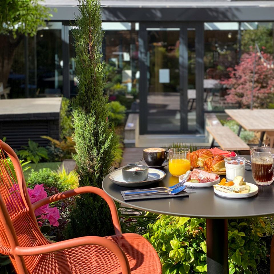 An orange chair a a small black table with breakfast on it, in the background is a green terrace