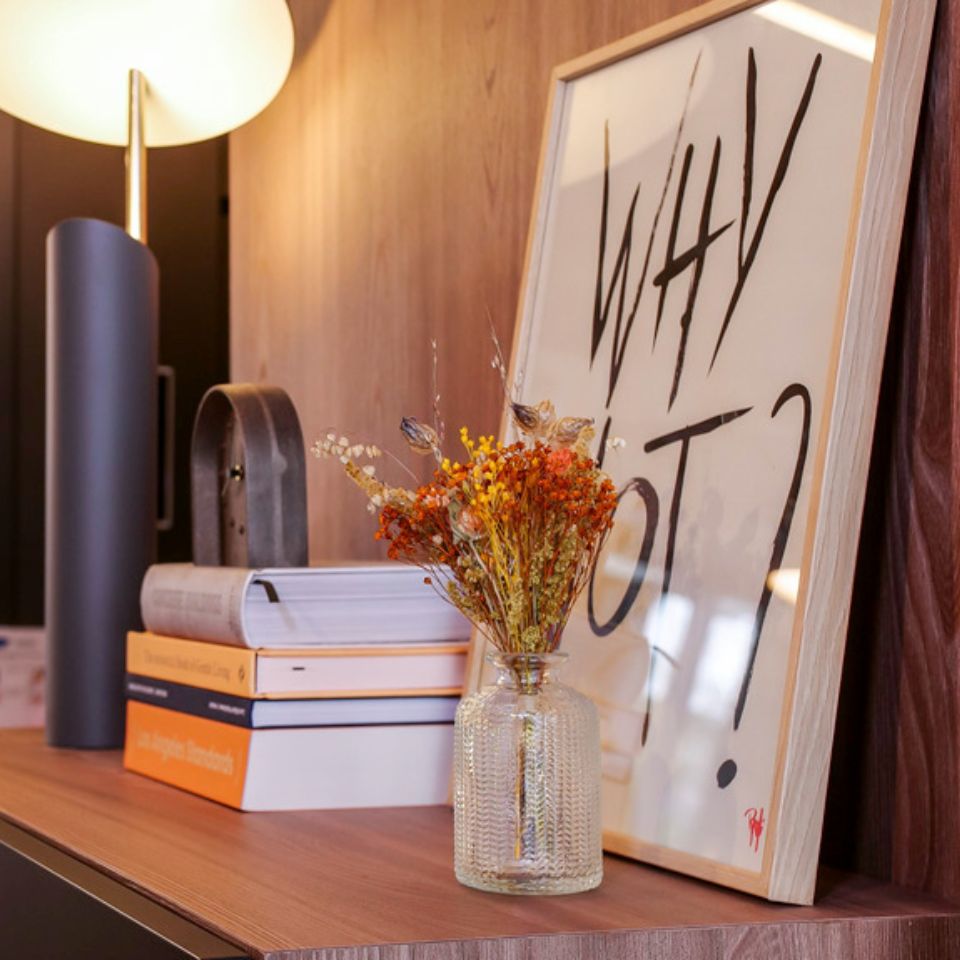A shelf with a stack of books, a framed picture and a glas vase with dried flowers
