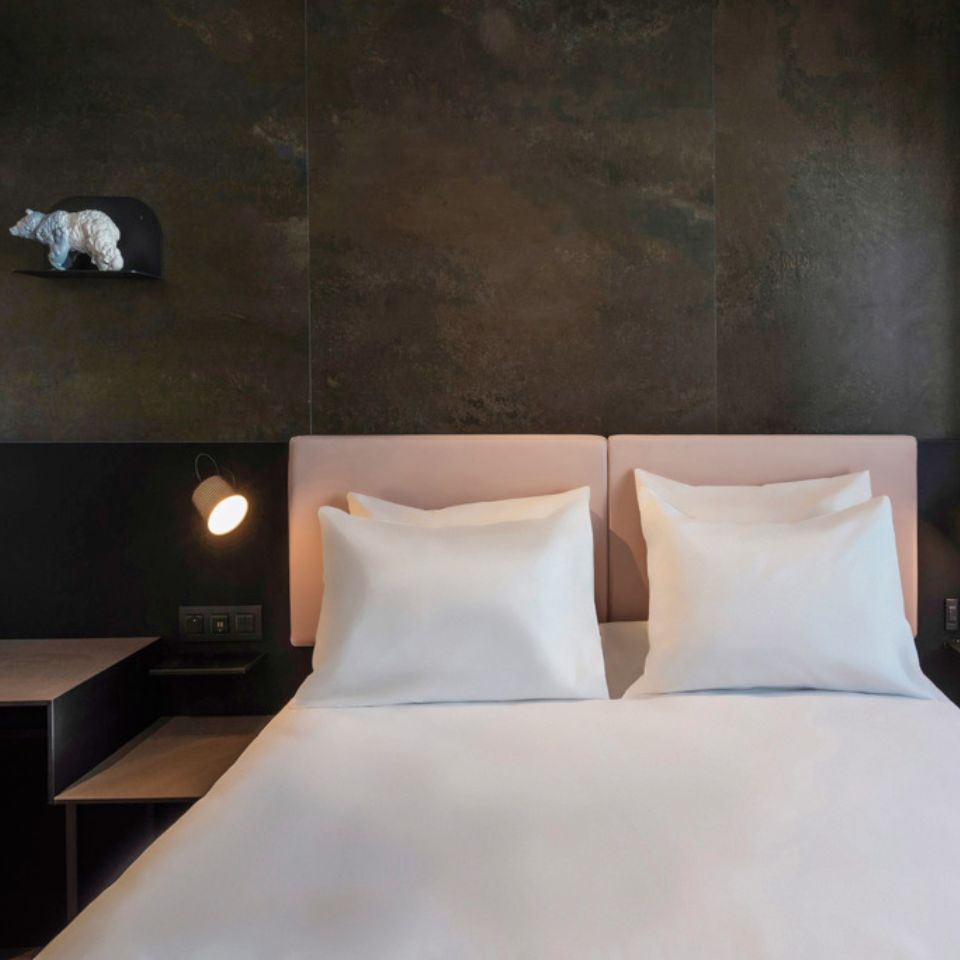 A guest room with a twin bed with a pink headboard in front of a black wall, on the left is a small figurine of a bear