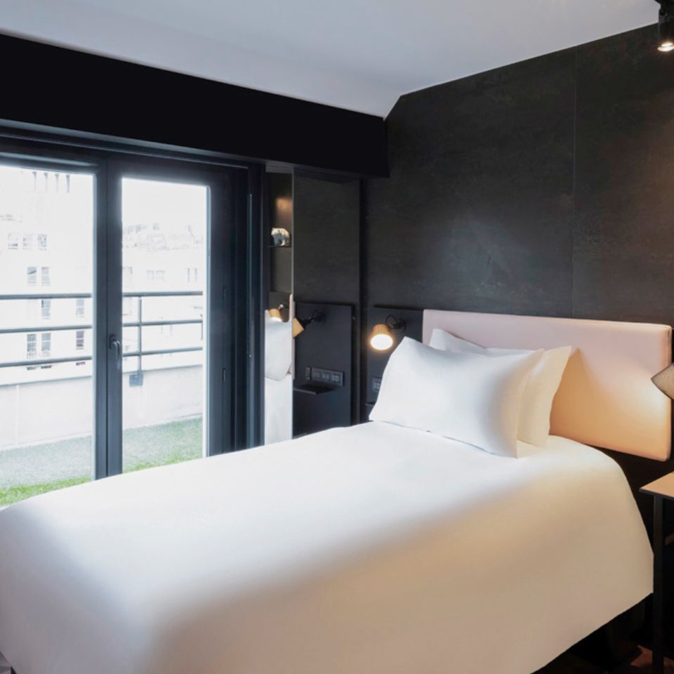 A single bed with white linens and a pink headboard in front of a window that opens into a terrace