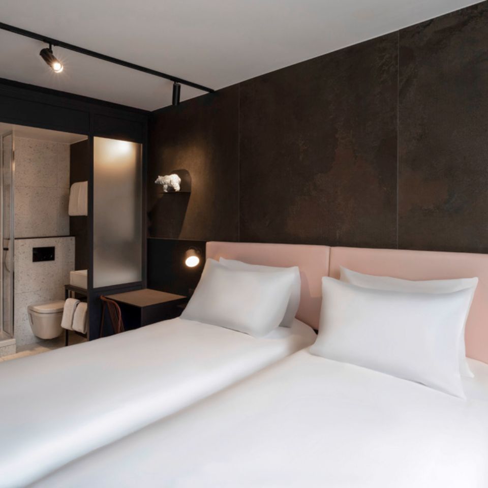 Twin beds with a pink headboard against a black wall. In the background is a bathroom